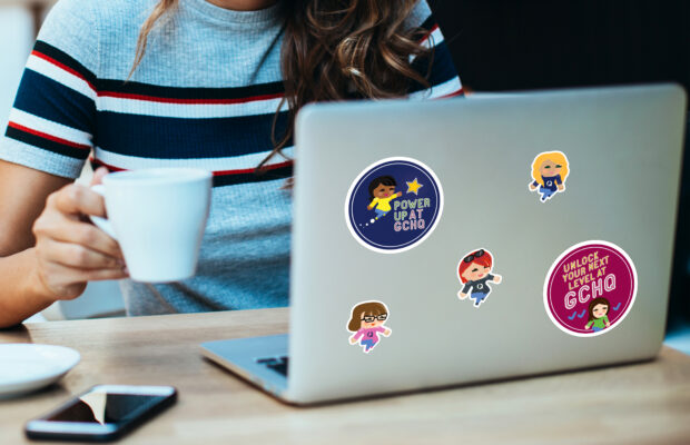 Image of a young woman with a cup of tea and a laptop featuring several stickers on its lid of graphics designed by Design102 in the Journey into the Known GCHQ recruitment campaign branding.