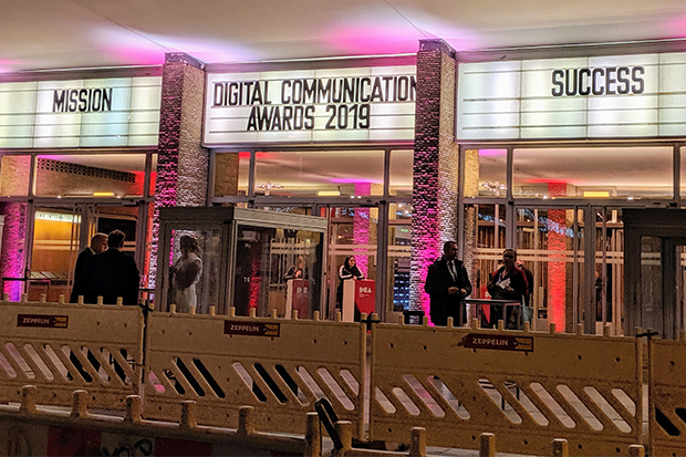 Evening exterior view of the cinema where the 2019 Digital Communications Awards ceremony was held in Berlin, Germany. A white clapperboard above the entrance doors is lit by pink spotlights and is divided into three sections. The centre section has letters spelling out the words Digital Communications Awards 2019. The sections either side display letters spelling out the words Mission and Success, relating to the outer space theme of the 2019 awards. Various people are standing outside the building talking.