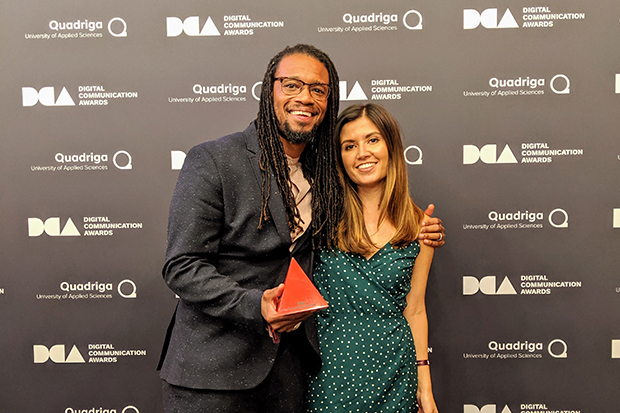 Holding the red triangular trophy as the award winners of the Digital Communications Awards 2019 Recruiting and Employer Branding category, Design102 team members Craig Ewing-Wilson - wearing a grey suit, and Tanya Hutnik - wearing a green sleeveless polka dot dress, stand in front of the media board listing the event logos. The logos for Digital Communications Awards and Quadriga University of Applied Sciences alternate left to right and top to bottom on the black background.