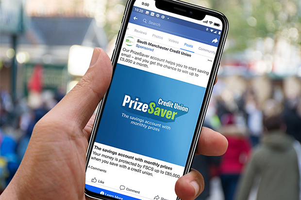 A photograph of a hand holding a smartphone showing a PrizeSaver Facebook advert. The Facebook caption reads: ‘Our PrizeSaver account helps you to start saving small - and you get the chance to win up to £5,000 a month’