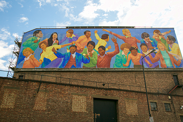 Mural painted on the side of a brick building. The mural depicts 18 children of different races running, playing and holding each other. The background is bright blue and their clothing is colourful: purple, yellow, red and orange. 