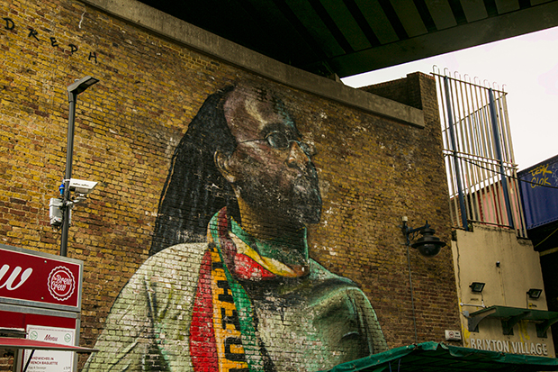 Mural painted on a brick wall beneath a railway bridge. The mural depicts Michael Johns, a local resident. Johns has long dreadlocks and small round glasses. In the painting he looks calmly upward while wearing a green t-shirt and red, yellow and green scarf. 