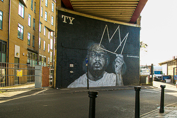 Mural painted on a brick wall at street level, beneath a railway bridge. The mural depicts the rapper TY, who died in 2020. It is a black-and-white painting. TY has his eyes closed and holds the outline of a crown in one hand. 