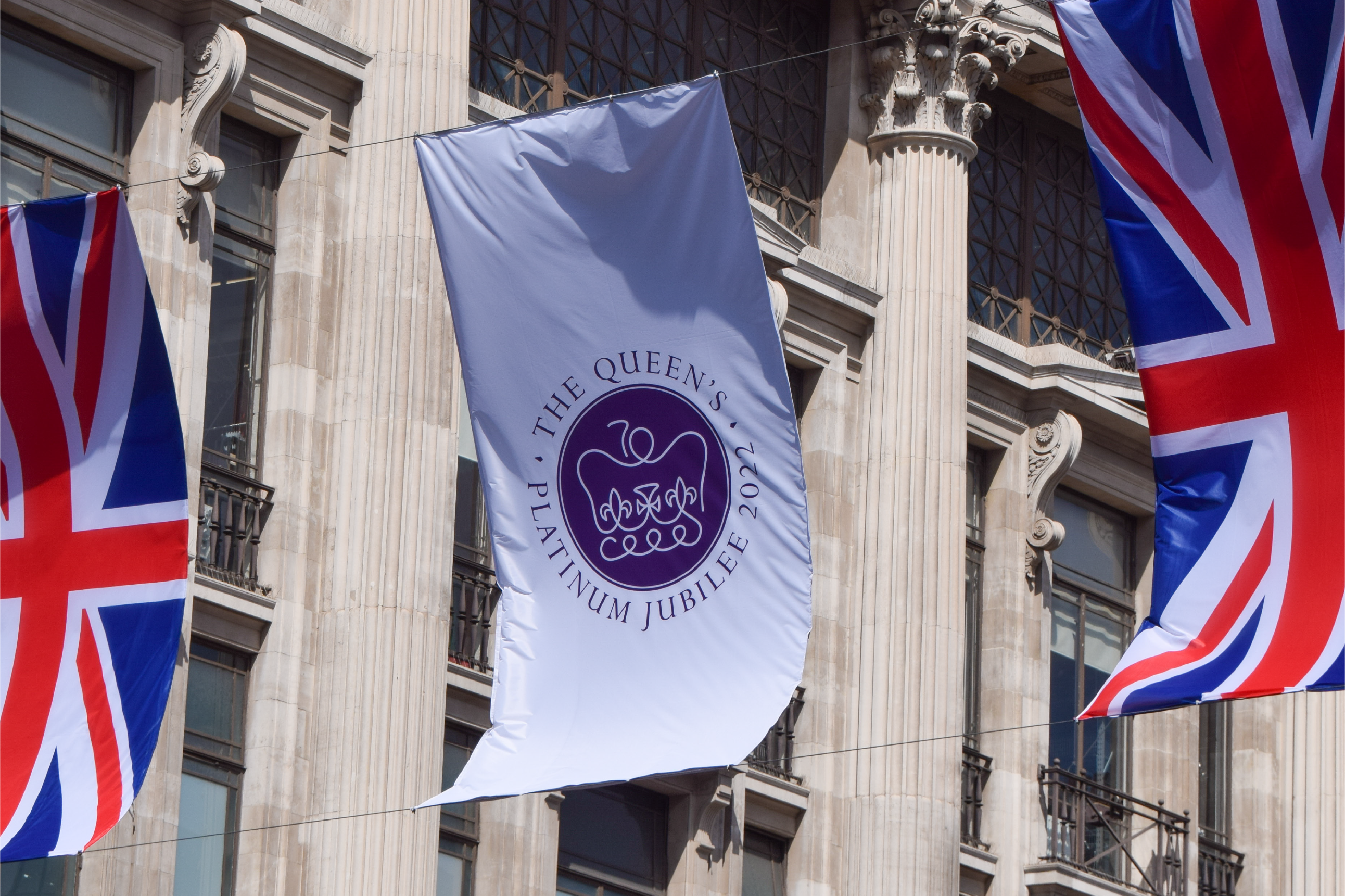 Three banners hanging over a London street. The middle one bears the purple Platinum Jubilee logo, either side of it are Union Jacks.