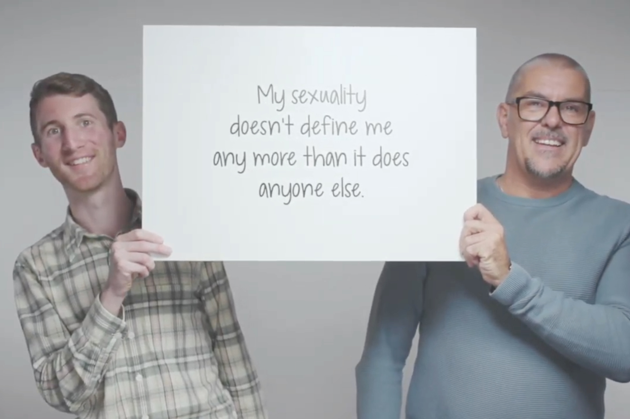 Still from a video featuring two smiling, adult men either side of a large white card they’re holding up between them. The card features the text “My sexuality doesn’t definite any more than it does anyone else”.