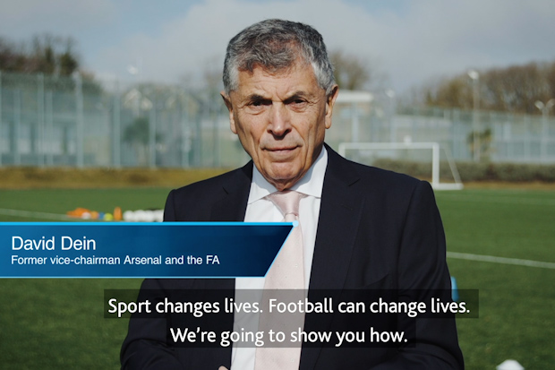 Screenshot of a video featuring a man speaking at a local football pitch-side interview. A name card indicates he is David Dein, Former vice-chairman Arsenal and the FA. Closed captions overlaying the image detail the dialogue: “Sport changes lives. Football can change lives. We're going to show you how.”