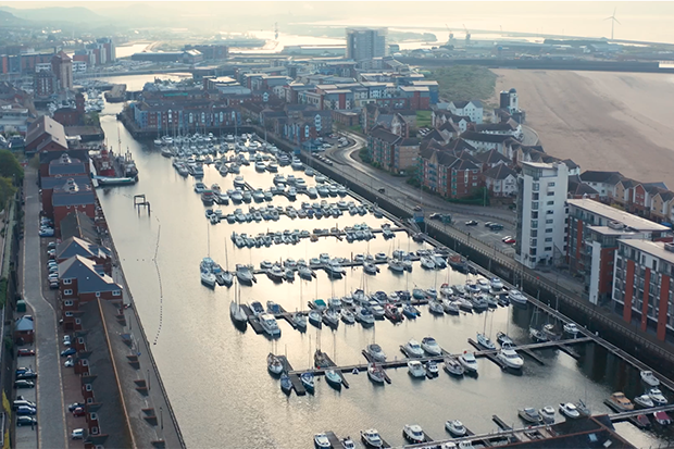 Aerial view of a marina taken by a drone, showing numerous boats docked along piers in a narrow waterway. In the background, a sandy beach, wind turbines, and an industrial port area are visible. This was captured as part of a video project for the Independent Monitoring Authority.